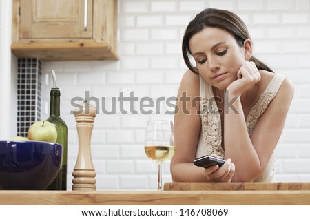 Young woman text messaging through cellphone while leaning on wooden counter in kitchen