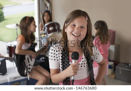 Portrait of cute young girl singing into microphone with friends playing musical instrument in garage