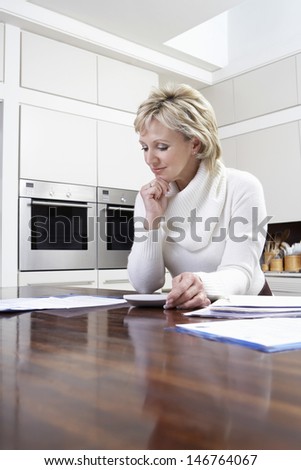 Middle aged woman calculating domestic bills with calculator in kitchen