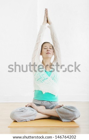 Full length of woman sitting cross legged with joined hands over head at gym