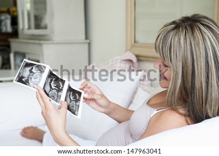 Happy young pregnant woman looking at baby scan on sofa