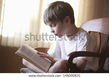 Young boy reading book on chair at home