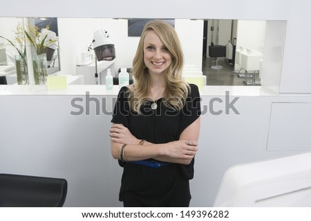Portrait of happy young hairstylist standing arms crossed at reception in salon