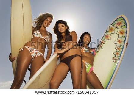 Low angle portrait of young friends in bikinis carrying surfboards against sky