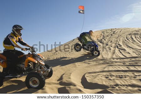 Side view of young men riding quad bikes over sand dunes in desert