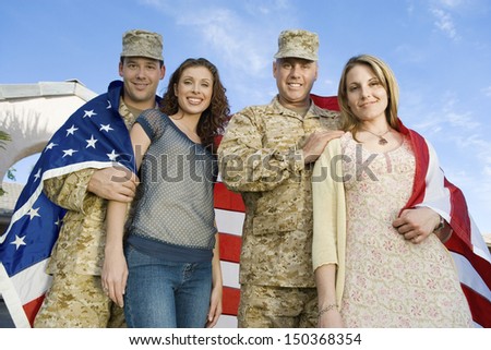 Low angle portrait of happy military couples wrapped in American flag against sky