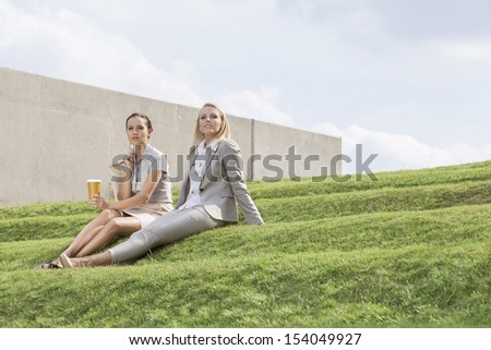 Full length of relaxed businesswomen in formals sitting on grass steps against sky
