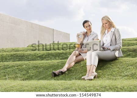 Full length of serious businesswomen looking at laptop while sitting on grass steps against sky