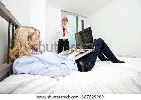 Businesswoman using laptop in bed with businessman adjusting tie at hotel