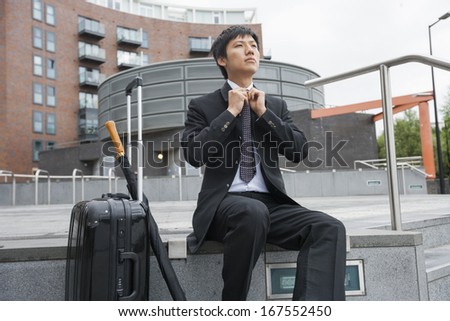 Mid adult businessman with luggage adjusting necktie against buildings