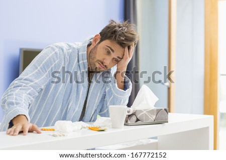 Young ill man with coffee mug; medicine and tissue leaning on kitchen counter