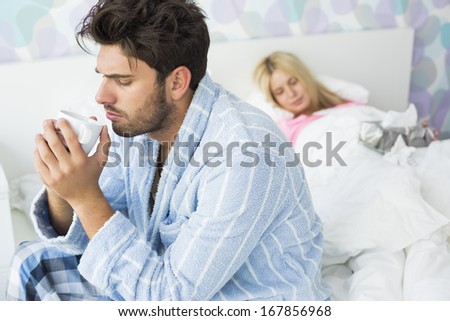 Sick man drinking coffee on bed while woman sleeping in background at home