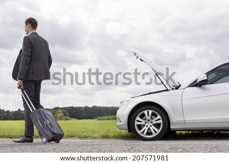 Full length rear view of young businessman with luggage leaving broken down car at countryside