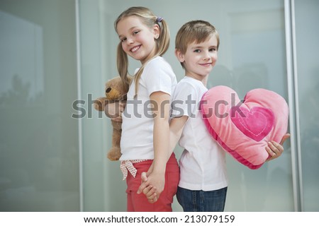 Boy and girl with heart shape cushion and teddy bear holding hands while standing back to back