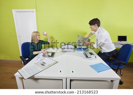 Young businesswoman throwing paper ball at male colleague holding waste bin