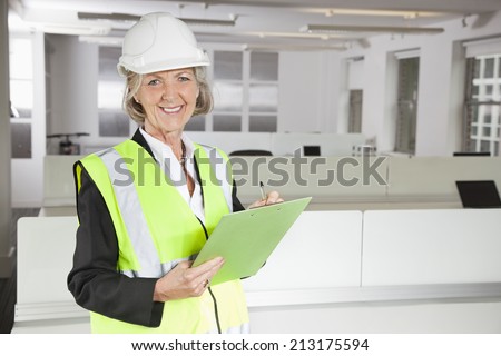 Portrait of smiling senior woman in reflector vest and hard hat holding clipboard at office
