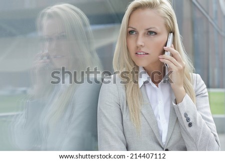 Beautiful young businesswoman conversing on cell phone while looking away by glass wall