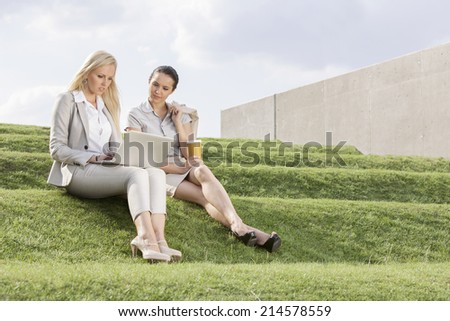 Full length of serious businesswomen looking at laptop while sitting on grass steps against sky