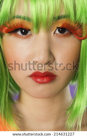 Close-up portrait of young woman wearing green wig and orange eyelashes over gray background
