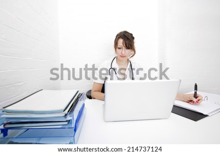 Young female doctor with laptop writing notes on desk in clinic