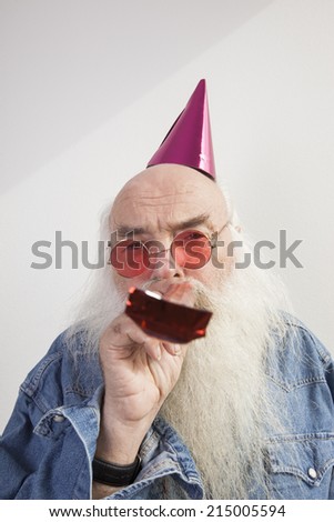 Portrait of senior man wearing party hat and red glasses while blowing horn against gray background