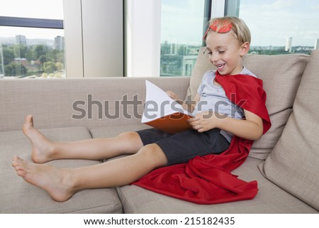 Happy little boy in superhero costume reading book on sofa at home