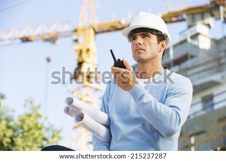Male architect with blueprints using walkie-talkie at construction site