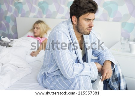 Sick man sitting on bed with woman sleeping in background at home
