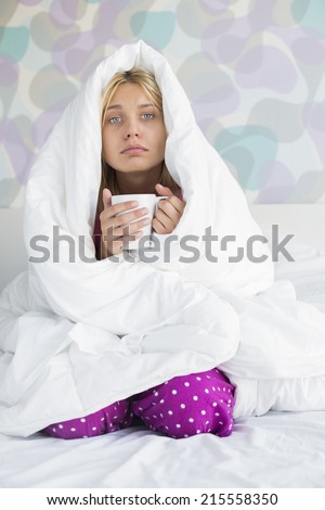 Portrait of young woman with coffee mug suffering from fever while wrapped in quilt on bed