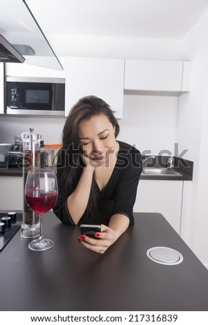 Happy young woman reading text message on smart phone in kitchen