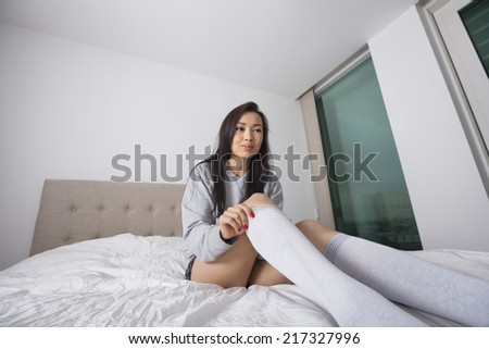 Thoughtful young woman putting on knee socks on bed