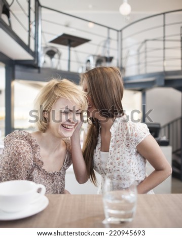 Happy young woman whispering into female friend\'s ear in cafe
