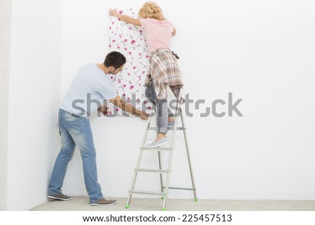 Full-length rear view of couple applying wallpaper to wall