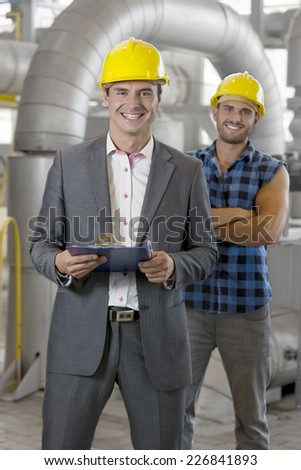 Portrait of smiling young male manager holding clipboard with worker in background at industry