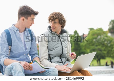 Young male college friends with laptop studying together in park