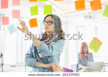 Creative businesswoman reading sticky notes on glass wall with colleague working in background at office
