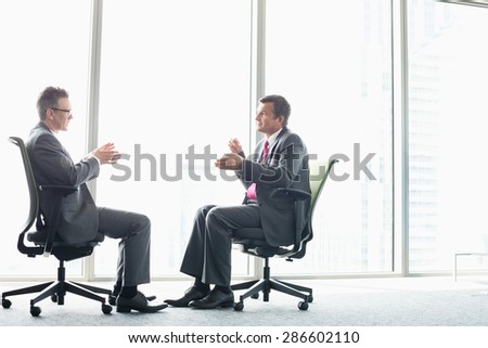 Full-length side view of businessmen discussing while sitting on office chairs by window