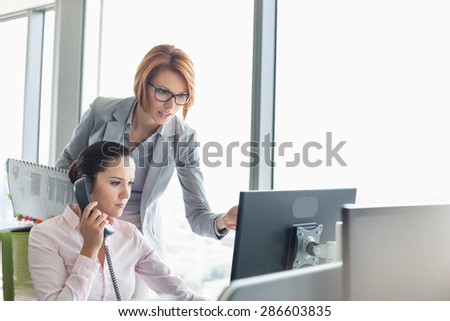 Young businesswoman using landline phone while colleague pointing at computer monitor in office
