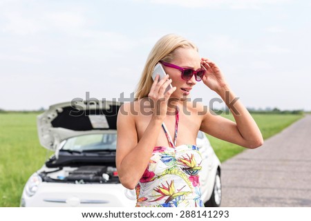 Tensed woman using cell phone on country road with broken down car in background