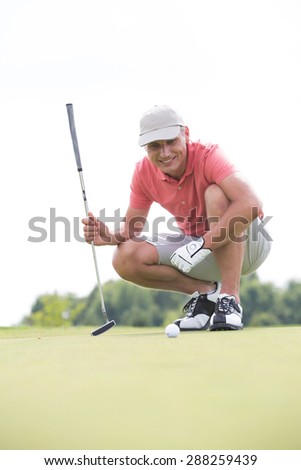 Smiling middle-aged man looking at ball while crouching on golf course