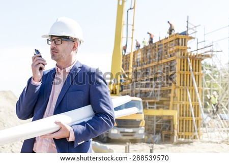 Architect using walkie-talkie while holding blueprints at construction site