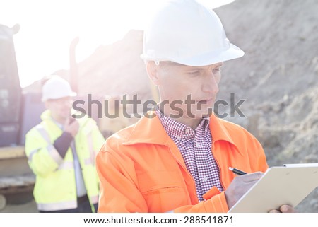 Engineer writing on clipboard at construction site with colleague in background