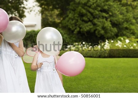 Two young girls in garden, wearing white dresses, holding balloons