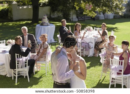 Mid adult bride and groom in garden among wedding guests, holding wineglasses, kissing