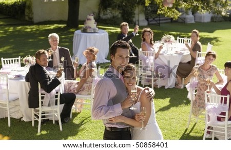 Mid adult bride and groom in garden among wedding guests, holding wineglasses, embracing
