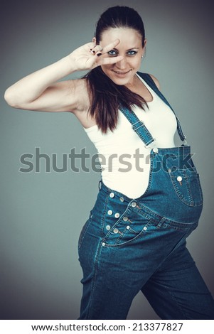 Pregnant beautiful girl in denim overalls. Pregnant woman posing on a gray background