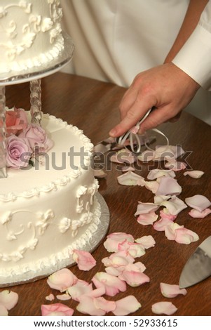 One hand cutting the wedding cake with a silver knife, flower petals on wood table in foreground.