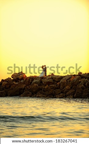 Bride and groom standing by the ocean on the rocks and looking at each other. Romantic moment at the sunset.