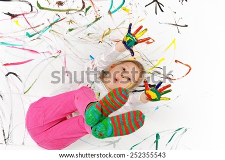 Little girl is playing with paints. She is painted all over and is lying on painted floor.