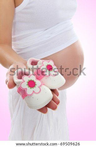 A pregnant woman mother holding baby shoes with belly exposed